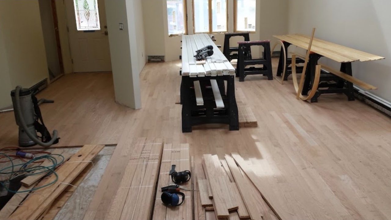 A Russotto installer hand-nailing a new hardwood floor in a great room with a stone fireplace, boards staged across the space